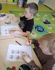 Dwoje uczniów siedzi przy ławkach rysując znaki zakazów palenia papierosów, na kartkach leżących przed nimi. Obok leżą przybory do rysowania, nożyczki i piórniki.