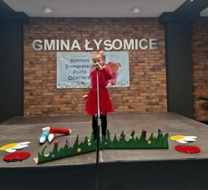 Uczennica oddziału przedszkolnego ubrana na czerwono z przyczepionymi na plecach skrzydłami motyla oraz w opasce z czerwonymi czułkami stoi przed mikrofonem na scenie. Przed dziewczynką widoczna jest wycięta z tektury trwa oraz biedronki, kwiaty i kredki ułożone na podłodze wokół niej. W tle widoczna jest ściana z cegieł z napisem "GMINA ŁYSOMICE" oraz umieszczonym pod nim jasnym banerem z widocznym napisem "Konkurs Interpretacji Poezji Dziecięcej".