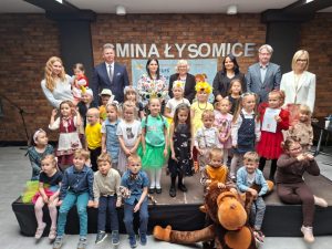 Uczestnicy konkursu wraz ze swoimi opiekunami i przedstawicielami z gminy pozują do wspólnego zdjęcia stojąc na ścianie wykonanej z cegieł z napisem "Gmina Łysomice". Jedna z dziewczynek ubrana na czerwono trzyma przed sobą otrzymany dyplom.