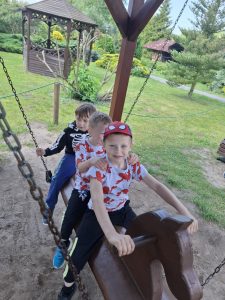 Trzech uczniów siedzi na drewnianej bujaczce w kształcie konia przymocowanej na łańcuch do drewnianej konstrukcji. W tle widoczny jest trawnik z drewnianą altaną.
