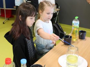 Dwie uczennice edukacji wczesnoszkolnej stoją przy stole. Jedna z nich trzyma w dłoni pipetę z niebieskim płynem ustawioną nad słoikiem z żółtą cieczą. Jej koleżanka przygląda się pipecie.