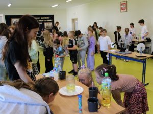 Uczniowie edukacji wczesnoszkolnej stoją rozproszeni na sali teatralnej, stawając przy różnych stanowiskach do wykonywania eksperymentów. Po środku widoczna jest kolejka uczniów oraz stolik z przyrządami do eksperymentów fizycznych. Na pierwszym planie widać dwie uczennice, które w skupieniu przyglądają się słoikowi z cieczą ustawionemu na stole przed nimi.