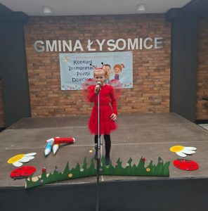 Uczennica oddziału przedszkolnego ubrana na czerwono z przyczepionymi na plecach skrzydłami motyla oraz w opasce z czerwonymi czułkami stoi przed mikrofonem na scenie. Przed dziewczynką widoczna jest wycięta z tektury trwa oraz biedronki, kwiaty i kredki ułożone na podłodze wokół niej. W tle widoczna jest ściana z cegieł z napisem "GMINA ŁYSOMICE" oraz umieszczonym pod nim jasnym banerem z widocznym napisem "Konkurs Interpretacji Poezji Dziecięcej".
