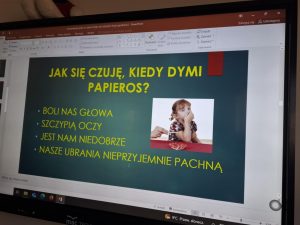 Zdjęcie przedstawia slajd z prezentacji multimedialnej wyświetlonej na monitorze komputera. Tytuł slajdu, zapisany dużymi żółtymi literami na ciemnym tle, brzmi: „Jak się czuję, kiedy dymi papieros?”. Pod tytułem znajduje się lista objawów: Boli nas głowa Szczypią oczy Jest nam niedobrze Nasze ubrania nieprzyjemnie pachną Po prawej stronie slajdu znajduje się zdjęcie małej dziewczynki, która siedzi przy stole i zasłania nos dłonią. Obok niej widać dorosłą dłoń trzymającą papierosa. Prezentacja znajduje się w programie PowerPoint, a na pasku dolnym ekranu widnieje informacja o pogodzie: „9°C, Przew. słonecz.” oraz ikony systemowe.
