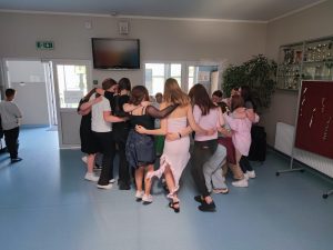 Grupa dzieci i młodzieży w ścisłym kręgu, przytulają się nawzajem w centralnej części szkolnego korytarza. Uczestnicy stoją blisko siebie w geście jedności lub zakończenia wspólnej aktywności. W tle otwarte drzwi prowadzą na zewnątrz, a po prawej stronie widoczna jest gablota z pucharami i roślina doniczkowa.