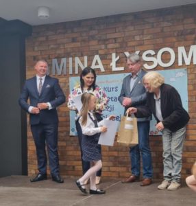 Uczennica biorąca udział w konkursie odbiera dyplom oraz nagrodę, od Pani reprezentującej organizatorów konkursu. Obok stoi trzech innych do rosłych, a w tle widoczna jest ściana z cegieł z niepełnym napisem "GMINA ŁYSOMICE".