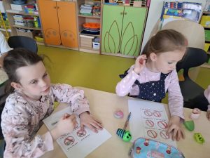 Dwie uczennice siedzą przy wspólnym stole. Przed dziewczynkami leżą kartki z narysowanymi odręcznie znakami zakazu palenia Jedna z dziewczynek trzyma w dłoni kredkę, druga sięga dłonią za ucho. Na stole leżą przybory do rysowania.