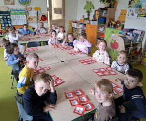 Uczniowie oddziału przedszkolnego siedzą przy dwóch dużych stołach. Dzieci patrzą w stronę obiektywu. Przed nimi leżą odręcznie wykonane znaki zakazu palenia przyklejone na czerwonych kwadratowych kartkach papieru.
