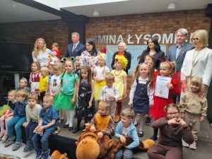 Uczestnicy konkursu wraz ze swoimi opiekunami i przedstawicielami z gminy pozują do wspólnego zdjęcia stojąc na ścianie wykonanej z cegieł z napisem "Gmina Łysomice". Jedna z dziewczynek ubrana na czerwono trzyma przed sobą otrzymany dyplom.