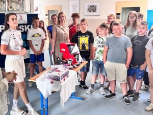 Uczniowie szkoły stoją zgromadzeni przy stoliku, na którym ustawione są nagrody oraz pudełko do losowania zwycięzców w konkursie. Za stolikiem stoi kobieta ubrana na sportowo.