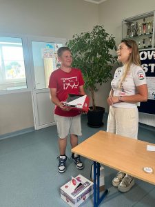 Uczeń klasy siódmej trzyma w dłoniach pudełko z kaskiem, który wygrał w konkursie. Obok niego stoi kobieta ubrana na sportowo. Przed nią widoczna jest ławka, przy której na podłodze leży karton obklejony logo gminy Łysomice. W tle widać fragment tablicy i gabloty z pucharami.