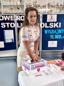 Kobieta ubrana na sportowo wkłada rękę do pudełka, w której znajdują się karteczki z nazwiskami uczniów biorących udział w konkursie. Za kobietą widoczna jest tablica z napisami "ROWEROWA STOLICA POLSKI", "WYGRAJMY 15 000 zł" ozdobiona grafikami postaci jeżdżących na rowerach.
