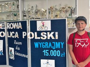 Uczeń z kaskiem rowerowym na głowie stoi obok tablicy z napisami "ROWEROWA STOLICA POLSKI", "WYGRAJMY 15 000 zł". "Razem dla Ostaszewa", która ozdobiona jest dodatkowo grafikami postaci jeżdżących na rowerach.