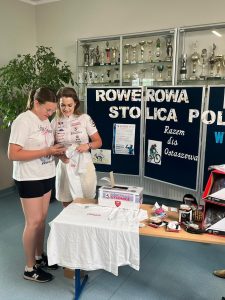 Uczennica szkoły stoi obok kobiety ubranej w sportowy strój. Obie patrzą na telefon, który dziewczynka trzyma w dłoni. Za nimi widoczna jest tablica z napisami "ROWEROWA STOLICA POLSKI", "Razem dla Ostaszewa" ozdobiona grafikami postaci jeżdżących na rowerach. Nad tablica widoczna jest gablota z pucharami zawieszona na ścianie.