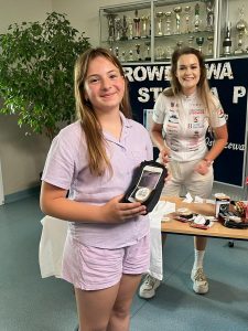 Uczennica klasy czwartej pozuje do zdjęcia trzymając w dłoniach przed sobą etui na telefon. Za nią widoczna jest ławka z pozostałymi nagrodami za którą stoi uśmiechnięta kobieta w stroju sportowym. W tle widoczny jest duży krzew oraz tablica z częściowo widocznymi napisem: "ROWEROWA STOLICA POLSKI". Nad tablicą wisi przytwierdzona do ściany gablota z pucharami.