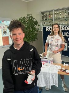 Uczeń klasy szóstej pozuje do zdjęcia trzymając w dłoniach małe pudełko zawierające wygraną przez niego nagrodę. Za nim widoczna jest ławka z pozostałymi nagrodami za którą stoi uśmiechnięta kobieta w stroju sportowym. W tle widoczny jest duży krzew oraz tablica z częściowo widocznymi napisem: "ROWEROWA STOLICA POLSKI". Nad tablicą wisi przytwierdzona do ściany gablota z pucharami.
