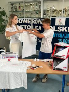 Uczeń klasy piątej stoi wraz z kobietą ubraną na sportowo za ławką na której rozłożone są nagrody w konkursie. Chłopiec wyciąga dłonie do trzymanej przez kobietę szeroko rozłożonej koszulki. W tle stoi tablica na której częściowo widoczne są napisy "ROWEROWA STOLICA POLSKI", "WYGRAJMY 15.000 ZŁ". Za tablicą na ścianie wisi gablota z pucharami.