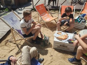 Czworo uczniów siedzi na leżakach na plaży. Po środku przed nimi stoi stolik na którym leży pizza. Dwoje z uczniów, którzy są w pełni widoczni w kadrze je pizze.