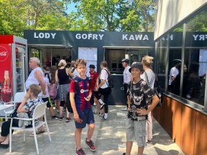 Uczniowie biorący udział w rajdzie stoją przed pomalowaną na czarno budką z napisem "LODY GOFRY KAWA". Dwoje uczniów odwraca sie i uśmiecha do zdjęcia.