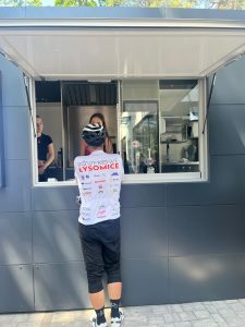 Przed budką z lodami stoi mężczyzna odwrócony plecami do obiektywu. Ubrany jest na sportowo, na głowie ma kask a na jego koszulce widoczny jest czerwony napis "Łysomice".