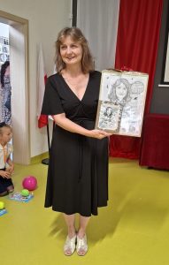 Wychowawczyni oddziału przedszkolnego pozuje do zdjęcia trzymając w dłoniach swoją karykaturę, którą otrzymała w prezencie.
