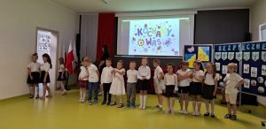Uczniowie oddziału przedszkolnego stoją w jednym rzędzie pozując do wspólnego zdjęcia. Jedna z dziewczynek odeszła ze swojego miejsca i stoi przy otwartych drzwiach prowadzących do sali. W tle ekran na którym widoczny jest kolorowy slajd z napisem "KOCHAMY WAS" oraz tablica z napisem "BEZPIECZNE WAKCJE".
