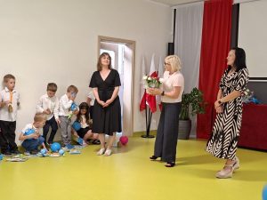 Na sali teatralnej stoi wychowawczyni oddziału przedszkolnego, pani dyrektor trzymająca bukiet kwiatów oraz jedna z mam uczniów. Dzieci stoją lub kucają pod ścianą oglądając otrzymane upominki.