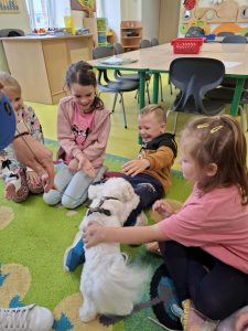 Uczniowie oddziału przedszkolnego siedzą na dywanie. Przed nimi stoi merdający ogonem mały, biały piesek. Jeden z chłopców wychyla się do tyłu ponieważ pies wspina się na jego nogi. Chłopiec jest szeroko uśmiechnięty i wyciąga swoją dłoń w kierunku psa.