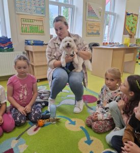 Kobieta siedzi na krześle ustawionym na dywanie w sali oddziału przedszkolnego. Trzyma w dłoniach małego białego pieska. Wokół niej na dywanie siedzą dzieci.