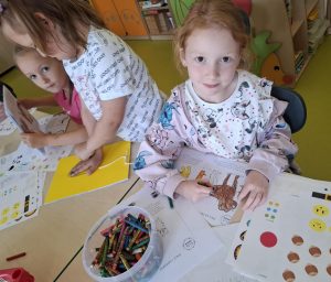 Trzy uczennice pracuje przy wspólnym stole wykonując prace plastyczne związane z psami. Na stole leżą kartki, arkusze z naklejkami i pudełko z kredkami świecowymi. Jedna z uczennic uśmiecha się delikatnie w stronę obiektywu. Dziewczynka koloruje kontur psa na kartce leżącej przed nią.