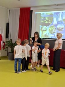 Grupa chłopców odbiera symboliczne upominki od wychowawczyni podczas zakończenia roku szkolnego na sali teatralnej. Obok stoi pani dyrektor. Jeden z chłopców trzyma w ręku tubę z dyplomem. W tle ekran z prezentacją oraz dekoracje w barwach narodowych.