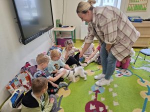Uczniowie oddziału przedszkolnego siedzą na dywanie w swojej sali. Dwóch chłopców głaszczę małego, białego pieska stojącego przed nimi. Nad pieskiem pochyla się z wyciągniętą ręką kobieta. Pani uśmiecha się szeroko.