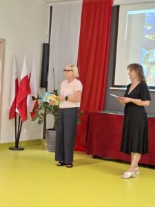 Wychowawczyni oddziału przedszkolnego i dyrektor szkoły stoją obok siebie trzymając w dłoniach książki. W tle widoczne są biało-czerwone flagi.