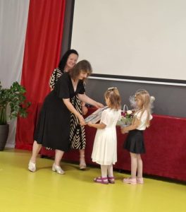 Wychowawczyni oddziału przedszkolnego odbiera od dwóch swoich uczennic upominki przygotowane na zakończenie roku szkolnego. Obok stoi uśmiechnięta mama jednego z dzieci. Po lewej stronie widoczna jest flaga polski.