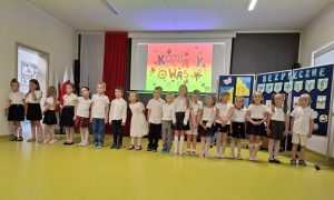 Uczniowie oddziału przedszkolnego stoją w jednym rzędzie pozując do wspólnego zdjęcia. W tle ekran na którym widoczny jest kolorowy slajd z napisem "KOCHAMY WAS" oraz tablica z napisem "BEZPIECZNE WAKCJE".