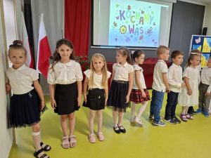 Grupa uczniów oddziału przedszkolnego stoi w jednym rzędzie pozując do wspólnego zdjęcia. Trzy dziewczynki patrzą wprost w stronę obiektywu. W tle ekran na którym widoczny jest kolorowy slajd z napisem "KOCHAMY WAS"