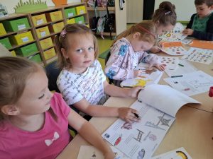 Pięcioro uczniów oddziału przedszkolnego siedzi przy wspólnym stole i wykonuje prace plastyczne dotyczące psów. Jedna z dziewczynek patrzy w stronę obiektywu opierając dłonie na blok a grafikami przedmiotów związanych z psami. Na stole przed uczniami leżą szablony z naklejkami oraz kredki.