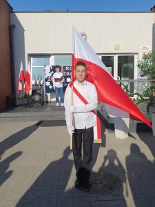 Uczeń trzymający oparta o ramię flagę stoi na wprost obiektywu na terenie przed budynkiem szkoły. Chłopiec ubrany jest w strój galowy, a przez ramię ma przewieszoną biało czerwoną szarfę.