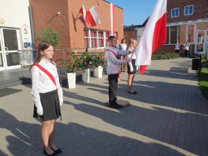 Na terenie przed budynkiem szkoły stoi troje uczniów w galowych strojach, którzy wchodzą w skład pocztu flagowego szkoły. Uczeń stojący po środku trzyma przed sobą flagę Polski. Uczniowie mają przewieszone przez ramiona biało czerwone szarfy, a na dłoniach białe rękawiczki.