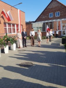 Przed budynkiem szkoły podczas inauguracji roku szkolnego stoi Pani dyrektor oraz troje przedstawicieli gminy Łysomice. Za nimi widoczny jest ustawiony bokiem poczet flagowy szkoły. Na budynku zamieszczone są polskie flagi.