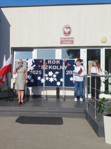 Przed wejściem do szkoły podstawowej stoi kobieta w sukience w biało czarną pepitkę, przemawia do mikrofonu. Obok niej dwoje uczniów odczytuje tekst z kartek. Za nimi widoczna dekoracja z napisem „Rok szkolny 2025/2026”. Po lewej stronie stoją dwie flagi Polski.