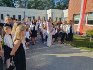 Grupa uczniów i nauczycieli zgromadzona na placu przed szkołą podczas inauguracji roku szkolnego. Dzieci i młodzież ubrane są odświętnie – w białe koszule, spódnice i ciemne spodnie. Obok nich stoją dorośli, również w eleganckich strojach.