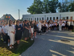 Grupa uczniów i nauczycieli zgromadzona na placu przed szkołą podczas inauguracji roku szkolnego. Dzieci i młodzież ubrane są odświętnie – w białe koszule, spódnice i ciemne spodnie. Obok nich stoją dorośli, również w eleganckich strojach.