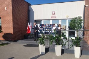 Przed wejściem do szkoły podstawowej stoi mężczyzna w garniturze, przemawia do mikrofonu. Obok niej dwoje uczniów odczytuje tekst z kartek. Za nimi widoczna dekoracja z napisem „Rok szkolny 2025/2026”. Po lewej stronie stoją dwie flagi Polski, a przed schodami ustawione są trzy białe donice z kwiatami.
