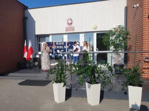 Przed wejściem do szkoły podstawowej stoi kobieta w kwiecistej sukni, przemawia do mikrofonu. Obok niej dwoje uczniów odczytuje tekst z kartek. Za nimi widoczna dekoracja z napisem „Rok szkolny 2025/2026”. Po lewej stronie stoją dwie flagi Polski.