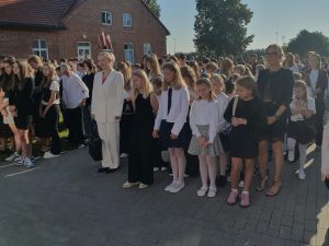Grupa uczniów i nauczycieli zgromadzona na placu przed szkołą podczas inauguracji roku szkolnego. Dzieci i młodzież ubrane są odświętnie – w białe koszule, spódnice i ciemne spodnie. Obok nich stoją dorośli, również w eleganckich strojach.