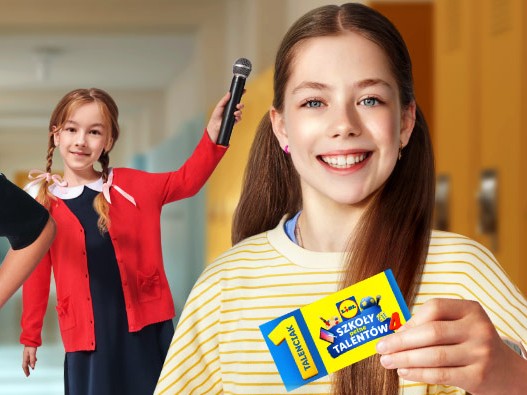 Uśmiechnięta szeroko dziewczynka widoczna na pierwszym planie trzyma w dłoni kupon z napisami: 1 talenciak, szkoły pełne talentów. Za nią widoczna jest dziewczynka w czerwonym sweterku trzymająca w wyciągniętej dłoni mikrofon.