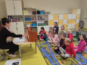 Grupa około dziesięciorga dzieci siedzi na kolorowym dywanie w sali szkolnej, zwrócona w stronę pani bibliotekarki. Kobieta siedzi przy małym stoliku i pokazuje ilustracje w teatrzyku kamishibai. W tle widać białe i żółte szafki oraz półki z książkami i materiałami dydaktycznymi.