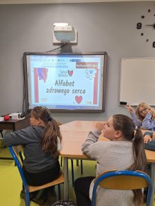 Grupa dzieci siedzi w sali lekcyjnej i patrzy na tablicę multimedialną, na której wyświetlona jest prezentacja „Alfabet zdrowego serca” z logotypem „Serce Dziecka”. Na pierwszym planie widać dwie dziewczynki siedzące tyłem, a w tle koleżanka pokazuje znak „V” palcami.