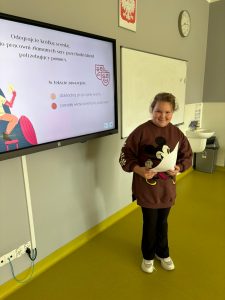 Dziewczynka w bordowej bluzie z Myszką Miki stoi przed klasą, trzyma kartkę i uśmiecha się. Obok na tablicy multimedialnej wyświetlony jest slajd z poleceniem: „Odegrajcie krótką scenkę, do pracowni złamanych serc przychodzi klient potrzebujący pomocy. W tekście zawrzyjcie: dokładną przyczynę wizyty, poradę właścicielki/a pracowni”.